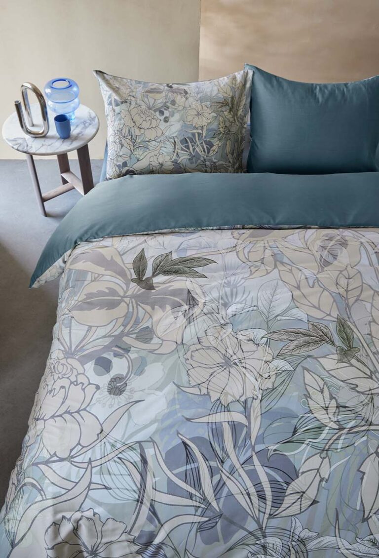 Ein Bett mit blauer, geblümter Bettwäsche von Beddinghouse Nobile steht neben einem kleinen runden Tisch, auf dem ein blaues Glas, eine Schale, ein Metallkrug und ein Buch in einer ruhigen, sanft beleuchteten Umgebung stehen.