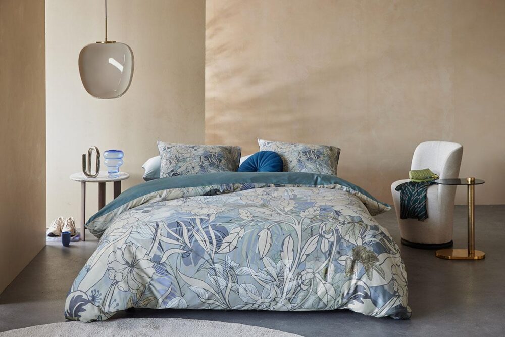 Ein modernes Schlafzimmer mit einem Bett aus Bettwäsche Beddinghouse Nobile, zwei gemusterten Kissen, einem tealfarbenen Kissen, einem runden Beistelltisch mit Dekor, einem weißen Sessel mit grüner Decke und einer Pendelleuchte vor beigen Wänden.