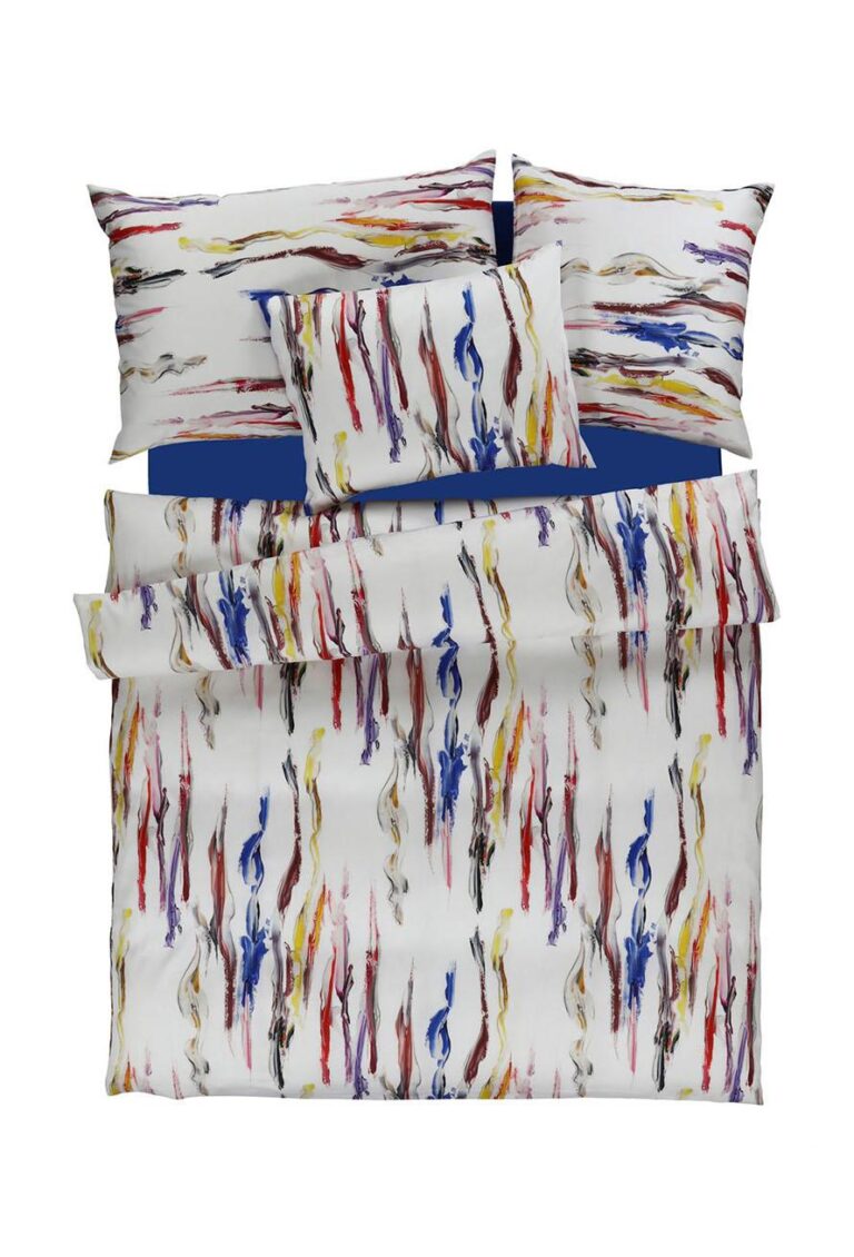 Ein Bett mit Bettwäsche Tamara-R Selection Alberto, mit weißen Laken, Kissenbezügen und einer Bettdecke mit bunten abstrakten Pinselstrichen in Blau, Rot, Gelb und Braun auf einem einfarbig blauen Bettlaken.