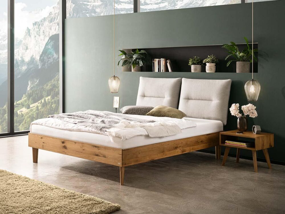 Modernes Schlafzimmer mit einem Holzbettgestell, weißer Bettwäsche und einem grauen Kopfteil. Es gibt Topfpflanzen und Dekoration auf einem Regal über dem Bett, Hängelampen, einen hölzernen Nachttisch und große Fenster mit Blick auf die Berge.
