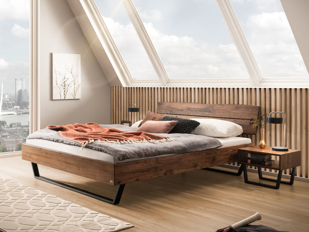 Ein modernes Schlafzimmer mit einem Holzbett, grauer und rosafarbener Bettwäsche, einem Nachttisch, einer Lampe und großen Dachfenstern mit Blick auf eine Stadtlandschaft. Neutrale Töne und natürliches Licht schaffen eine gemütliche, minimalistische Atmosphäre.