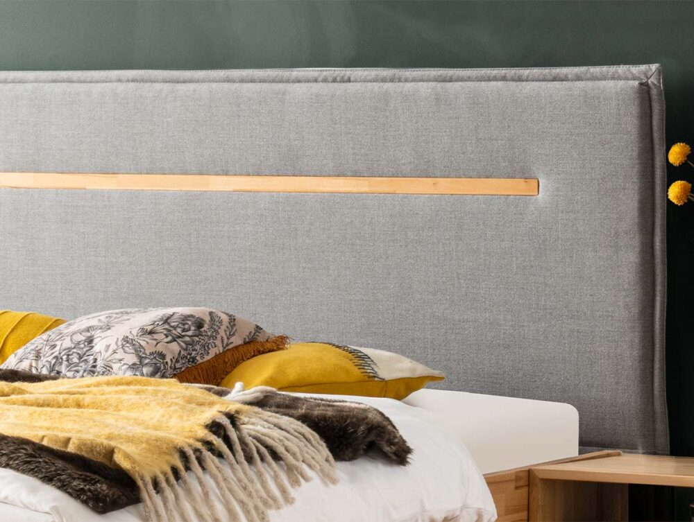 Ein ordentlich gemachtes Bett mit einem grau gepolsterten Kopfteil mit einem horizontalen Holzakzent. Das Bett hat gemusterte und gelbe Kissen und eine gelbe und braune Decke. Ein kleiner Nachttisch mit gelben Blumen ist sichtbar.