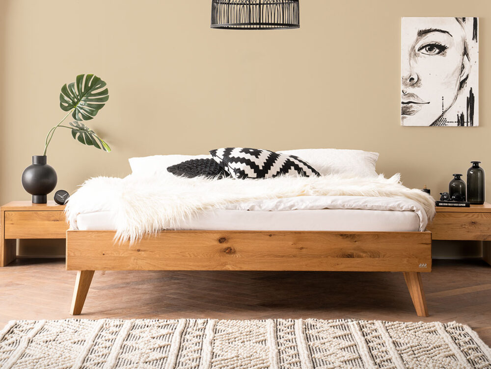 Ein gemütliches Schlafzimmer mit einem Holzbett, weißer Bettwäsche, einem schwarz-weiß gemusterten Kissen und einer flauschigen Decke. Es gibt Nachttische aus Holz, schwarze Dekoartikel, ein grünes Blatt in einer Vase und moderne Wandkunst über dem Bett an einer beigen Wand.