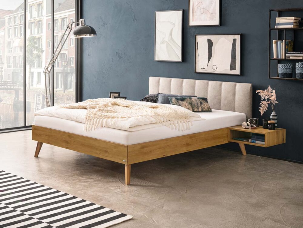 Ein modernes Schlafzimmer mit einem Bettgestell aus Holz, einem beigefarbenen gepolsterten Kopfteil und weißer Bettwäsche. Über dem Bett hängt Kunst an der Wand, und ein schwebender Nachttisch mit einer rosa Pflanze ist angebracht. Große Fenster und ein gestreifter Teppich vervollständigen den Raum.