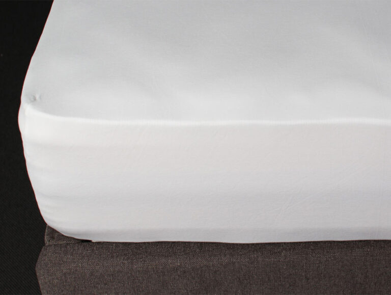 Nahaufnahme eines weißen Spannbettlakens, das die Ecke einer Matratze, die auf einem dunkelgrauen Bettgestell liegt, sauber abdeckt.