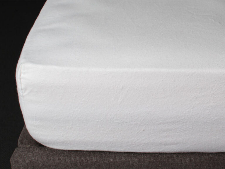 Nahaufnahme eines ordentlich gemachten Bettes, das die Ecke einer Matratze zeigt, die mit einem einfachen weißen Spannbettlaken bedeckt ist und auf einem dunklen Bettrahmen vor einem schwarzen Hintergrund ruht.