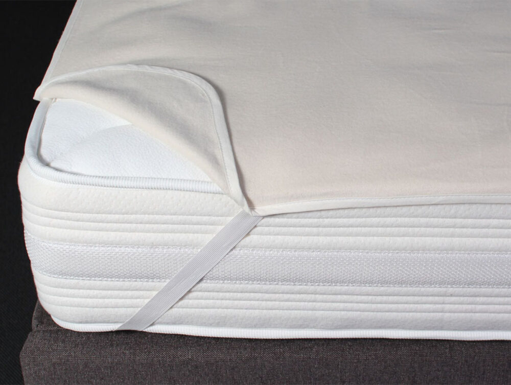 Nahaufnahme einer Matratzenecke mit einem teilweise zurückgezogenen Matratzenschoner, der mit elastischen Bändern über der Matratze auf einem dunklen gepolsterten Bettgestell befestigt ist.