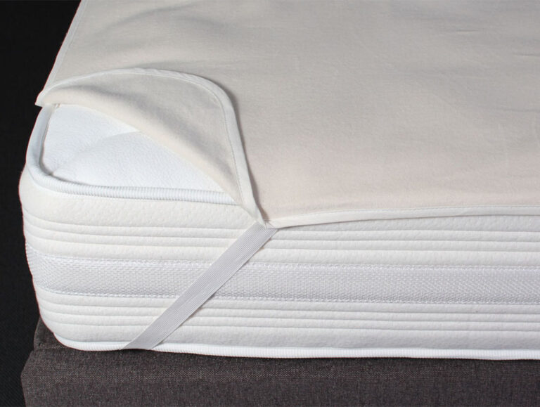 Nahaufnahme einer Matratzenecke mit einem teilweise zurückgezogenen Matratzenschoner, der mit elastischen Bändern über der Matratze auf einem dunklen gepolsterten Bettgestell befestigt ist.