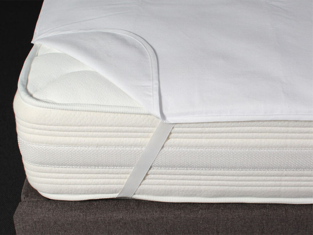 Eine Nahaufnahme einer weißen Matratze mit einem teilweise zurückgezogenen Matratzenschoner, der mit einem elastischen Band an der Ecke befestigt ist. Die Matratze liegt auf einem dunkelfarbigen Lattenrost.