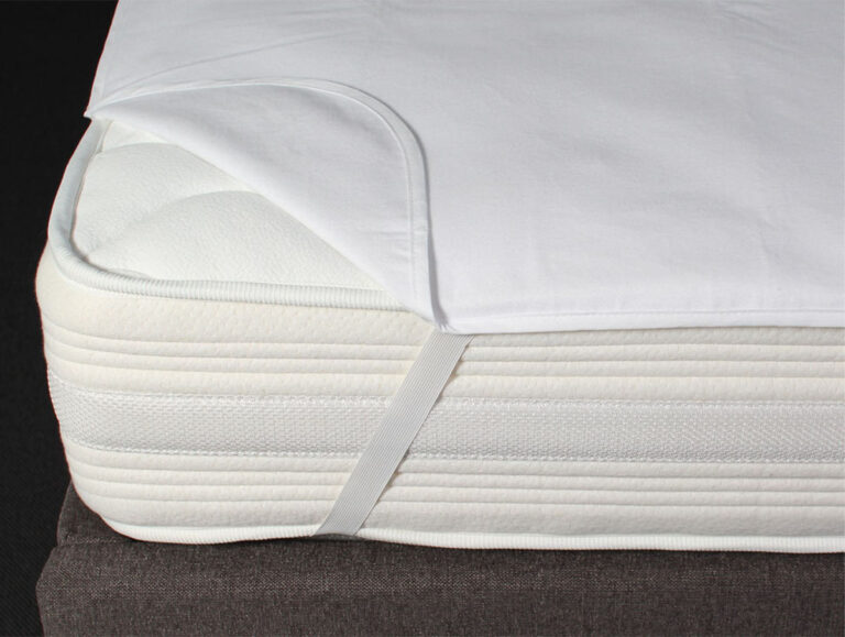 Eine Nahaufnahme einer weißen Matratze mit einem teilweise zurückgezogenen Matratzenschoner, der mit einem elastischen Band an der Ecke befestigt ist. Die Matratze liegt auf einem dunkelfarbigen Lattenrost.