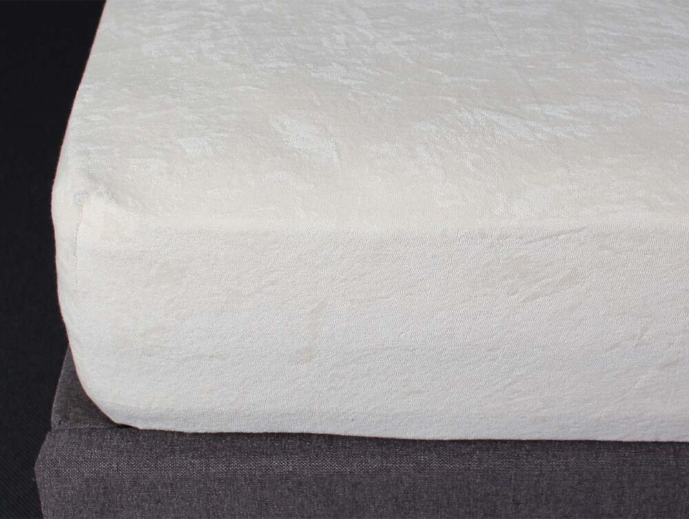 Nahaufnahme der weißen Ecke der Tamara-R Fix-Molton Classic Memory-Foam-Matratzenauflage auf einem dunkelgrauen Lattenrost, die die glatte, aber dezent strukturierte Oberfläche hervorhebt.