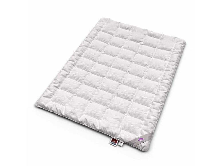 Eine weiße, rechteckige, gesteppte Matratzenauflage mit einem Gittermuster auf einem schlichten weißen Hintergrund. An einer Ecke der Matratzenauflage sind zwei Stoffanhänger zu sehen.