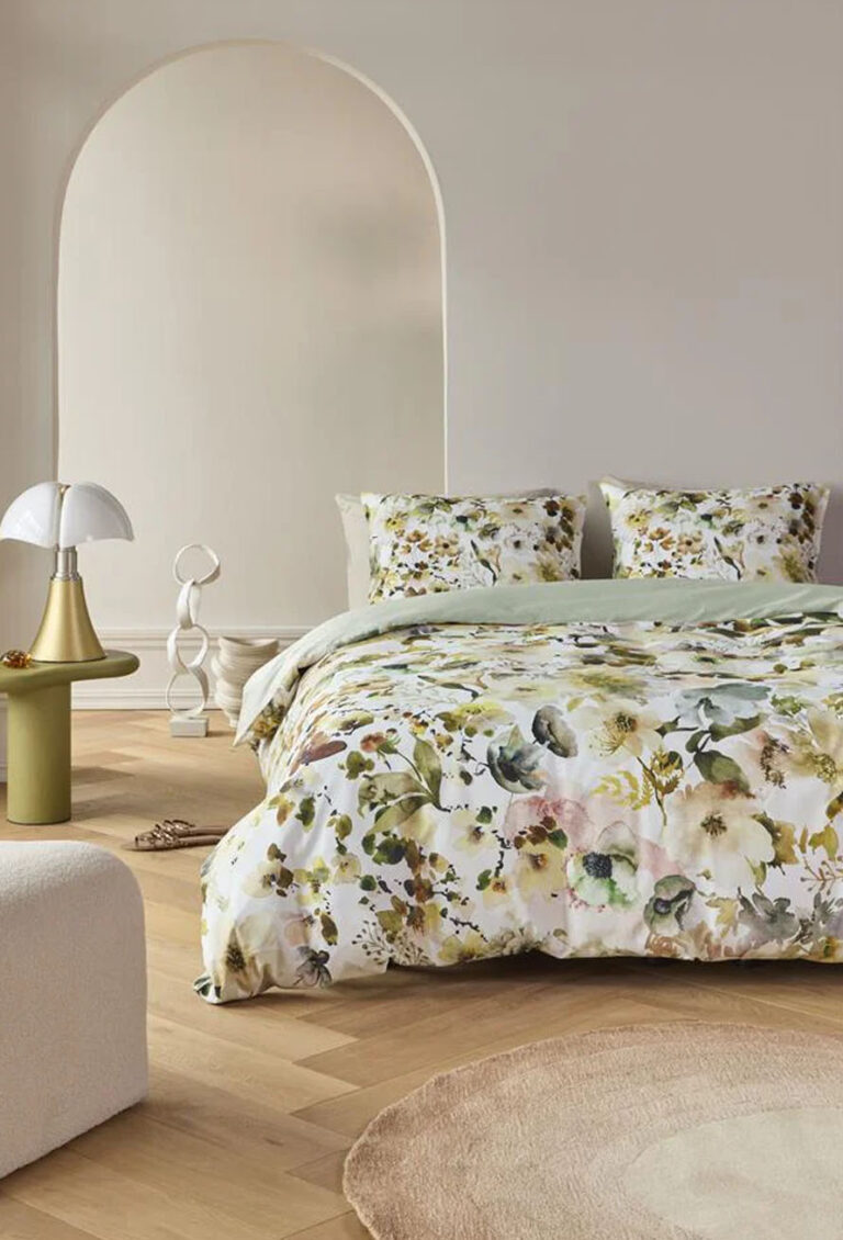 Ein modernes Schlafzimmer mit geblümter Bettwäsche von Beddinghouse Aquitaine, zwei passenden Kissen, hellen Holzböden, einem runden beigen Teppich, einem grünen Beistelltisch mit einer weißen Lampe und neutralen Wänden mit einer gewölbten Nische.