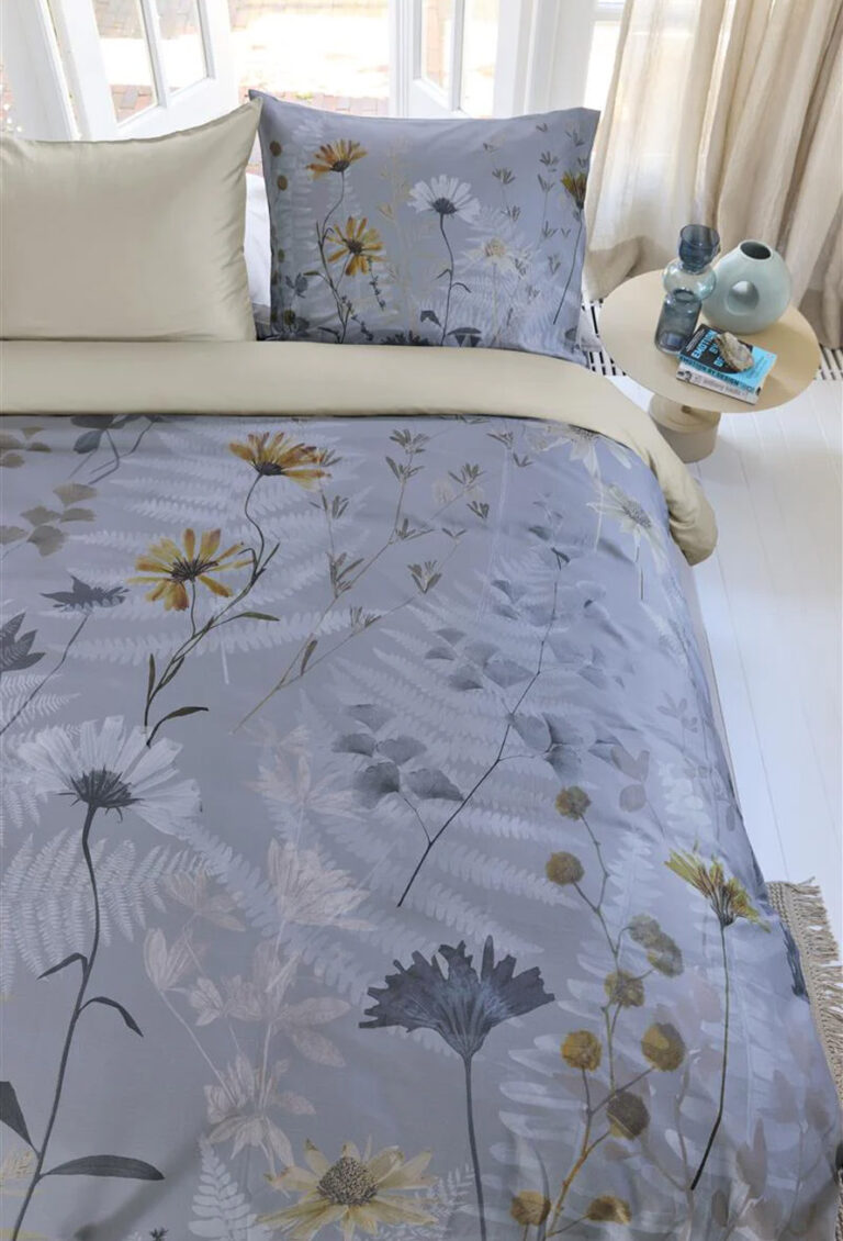 Ein ordentlich gemachtes Bett mit hellbeigen Laken und Bettwäsche von Bettwäsche Beddinghouse Nala mit Blumenmuster in Grau, Gelb und Weiß steht neben einem kleinen runden Tisch mit Vasen an sonnenbeschienenen Fenstern mit transparenten Vorhängen.