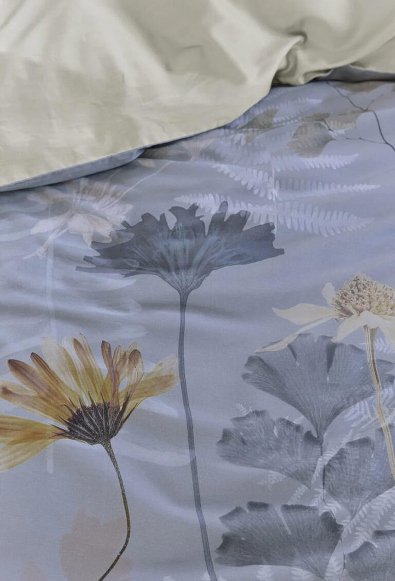 Nahaufnahme eines Bettes mit cremefarbenen Laken und dem Bettbezug Nala von Bettwäsche Beddinghouse, auf dem große graue, gelbe und weiße botanische Blumen und Farne auf hellblauem Hintergrund zu sehen sind.