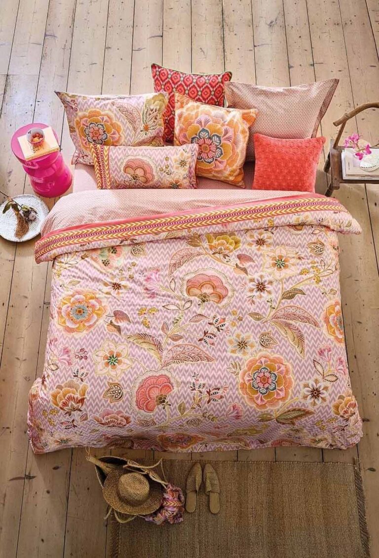 Ein gemütliches Bett ist mit Bettwäsche Pip Studio Hakuna bezogen, mit rosa, orange und gelben Blumenmustern und Kissen auf einem hellen Holzboden. Auf einem rosafarbenen Beistelltisch stehen neben einem Tablett mit einer Teekanne, einem Hut, Hausschuhen und einer geflochtenen Tasche weitere Gegenstände.