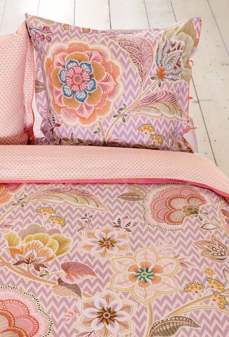 Ein Bett mit Bettwäsche Pip Studio Hakuna - eine rosa-weiße Zickzack-Bettdecke mit großen, farbenfrohen Blumendrucken in Rosa, Gelb, Orange und Blau - ruht auf einem hellen Holzboden.