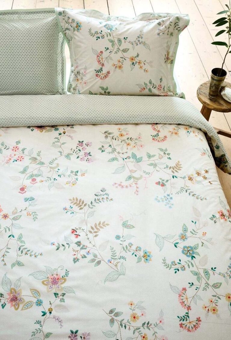 Ein ordentlich gemachtes Bett mit der weißen Bettwäsche Pip Studio Kili Flower und bunten Blumenkissen. Auf einem kleinen runden Nachttisch aus Holz stehen eine grüne Pflanze und eine Tasse, darunter befinden sich helle Holzdielen.