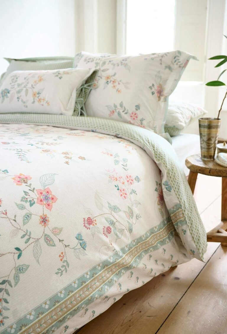 Ein ordentlich gemachtes Bett mit weißer Bettwäsche Pip Studio Kili Flower, mit einem pastellfarbenen Blumenmuster und grünen Akzenten, gepaart mit passenden Kissen und einer grünen Strickdecke. Ein Holzhocker mit einer Pflanze und einer Tasse steht neben dem Bett in einem hellen Raum.