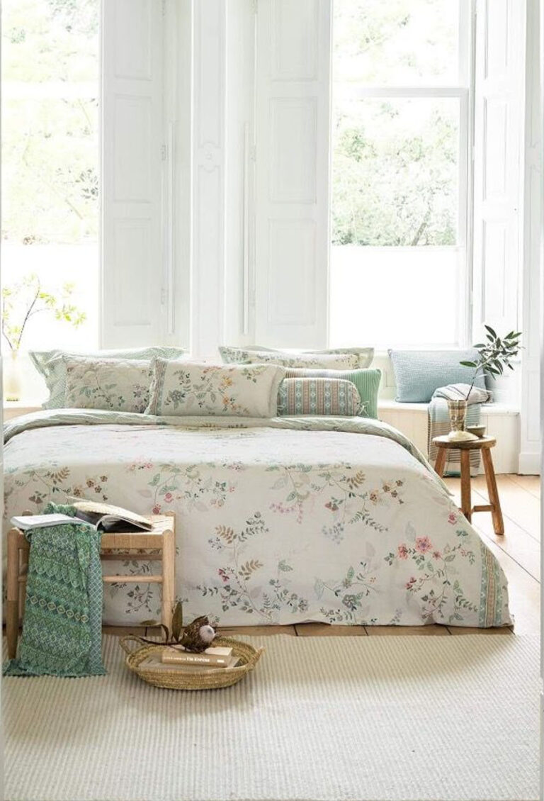 Ein helles Schlafzimmer mit großen Fenstern, weißen Wänden und einem Bett, das mit Bettwäsche Pip Studio Kili Flower bezogen ist. Helle Holzmöbel, ein Webteppich, eine kleine Pflanze auf einem Hocker und ein Korb mit Büchern und einer Tasse vervollständigen den Look.
