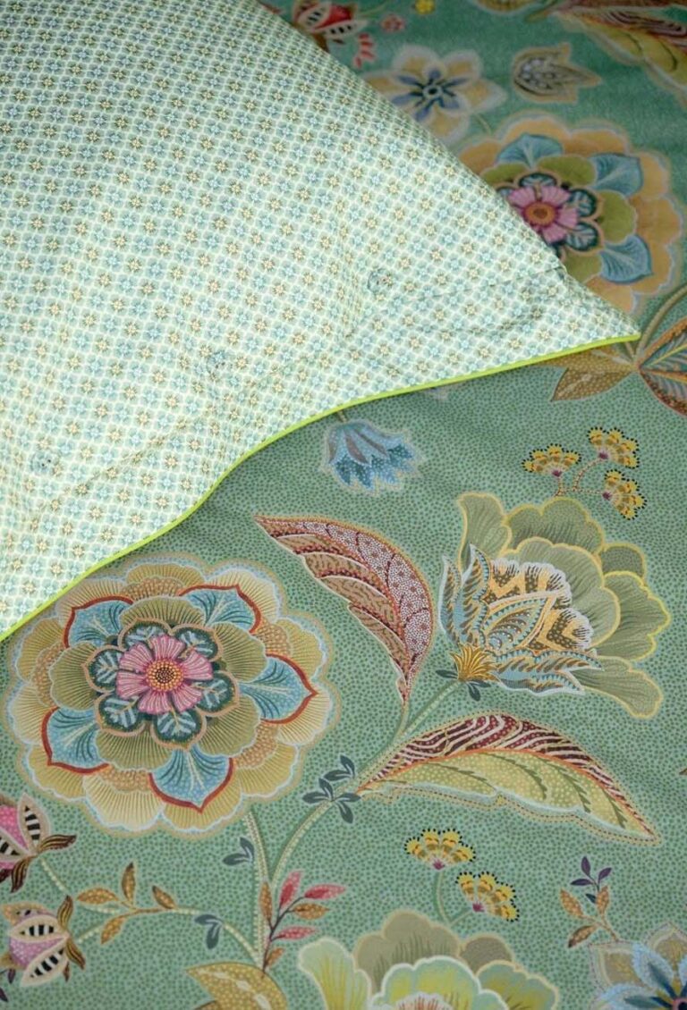 Eine Nahaufnahme eines Bettes zeigt Bettwäsche Pip Studio Matata: eine grüne, mit Blumen gemusterte Steppdecke und ein Kissen mit blauen und gelben geometrischen Mustern, die beide durch farbenfrohe, verschlungene Muster hervorgehoben werden.