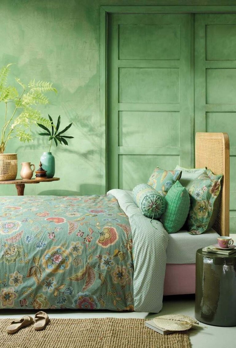 Ein gemütliches Schlafzimmer mit grünen Wänden und Türen, einem Kopfteil aus Rattan und einem Bett aus Bettwäsche Pip Studio Matata. Ein runder Beistelltisch hält eine Teetasse, während Pflanzen und Vasen die Holzkonsole schmücken.
