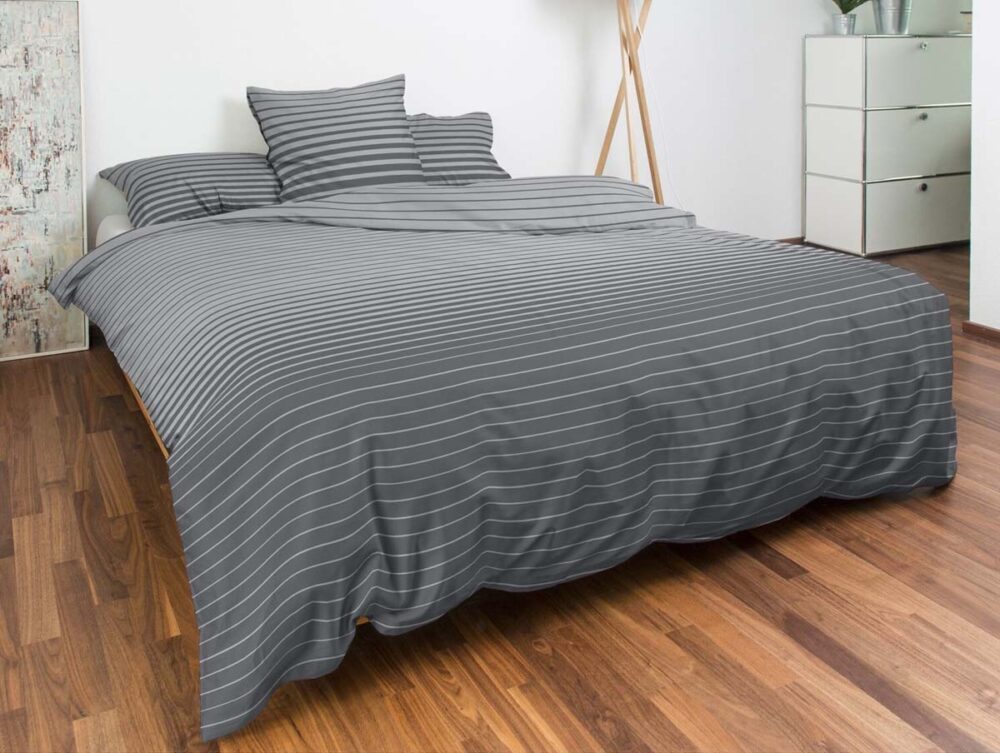 Ein ordentlich gemachtes Bett mit grau-weiß gestreifter Bettwäsche und Kissen von Bettwäsche Nobilium Weilo in einem modernen Schlafzimmer mit Holzböden, einer weißen Kommode und einer hohen Stehlampe in der Ecke.