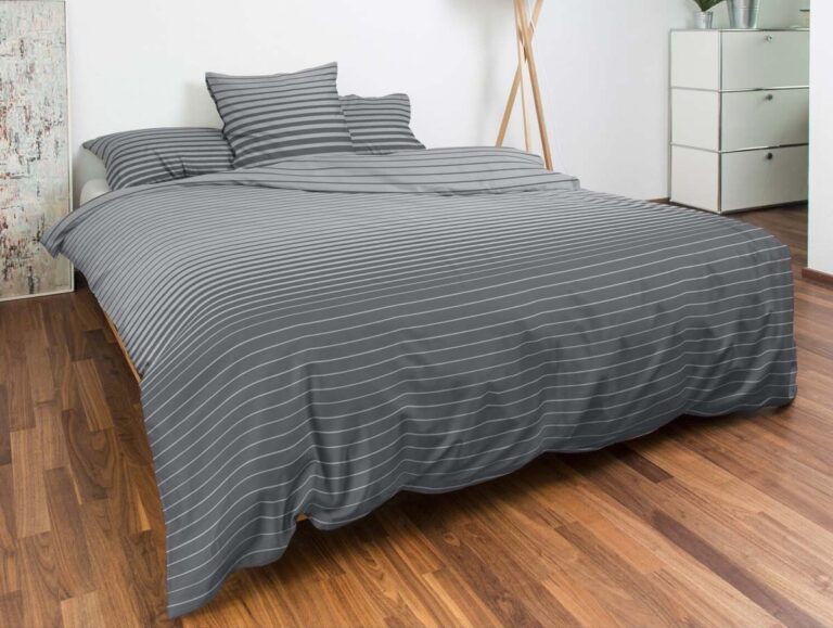 Ein ordentlich gemachtes Bett mit grau-weiß gestreifter Bettwäsche und Kissen von Bettwäsche Nobilium Weilo in einem modernen Schlafzimmer mit Holzböden, einer weißen Kommode und einer hohen Stehlampe in der Ecke.