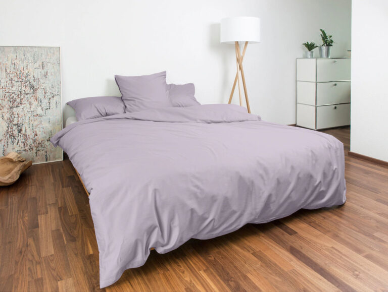 Ein ordentlich gemachtes Bett mit heller lavendelfarbener Bettwäsche steht in einem modernen Schlafzimmer mit Holzfußboden, einer weißen Stehlampe, Topfpflanzen, einer weißen Kommode und einem abstrakten Kunstwerk, das an der Wand lehnt.