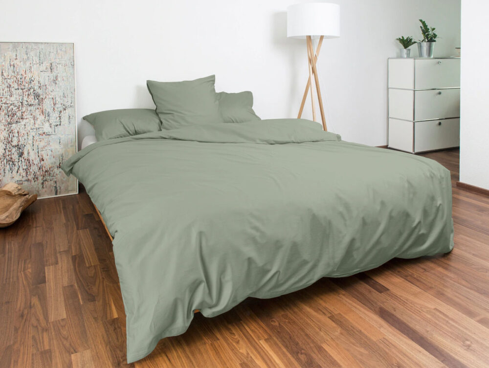 Ein modernes Schlafzimmer mit grüner Bettdecke und Kissen auf einem Doppelbett, Holzboden, weißen Wänden, einer Stehlampe, einer Kommode mit Pflanzen und einem strukturierten Kunstwerk, das an der Wand lehnt.