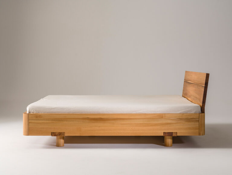 Ein minimalistisches Holzbett mit hellem Rahmen und einfachem Kopfteil, das sich durch klare Linien und ein niedriges Profil auszeichnet und vor einem schlichten weißen Hintergrund steht.