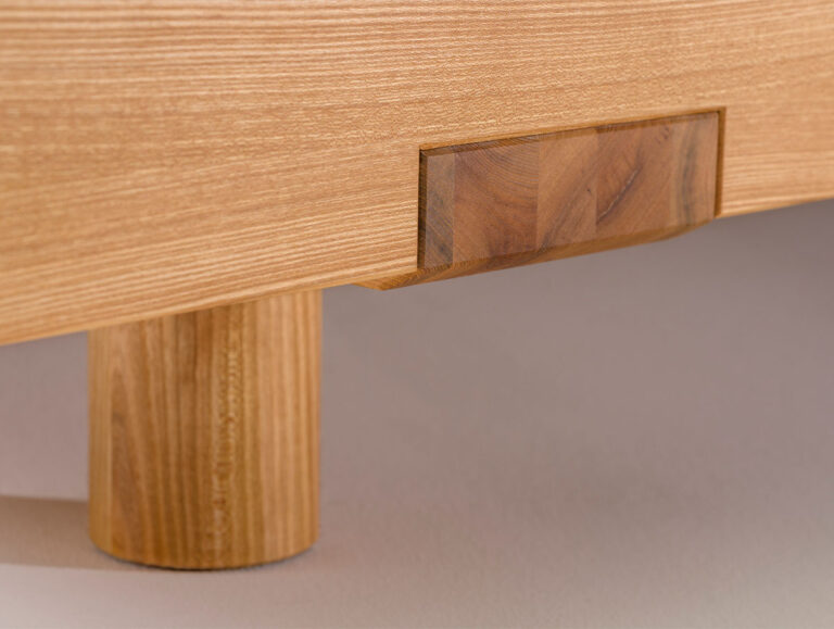 Nahaufnahme des Beins und der Kante eines Holztisches oder eines Möbelstücks, die die Holzmaserung und eine rechteckige dekorative Einlage zeigen. Die Oberfläche ist glatt und von heller Farbe.