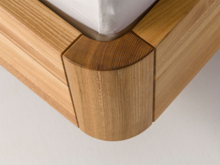 Nahaufnahme einer hölzernen Bettrahmenecke mit glatten, abgerundeten Kanten und heller Holzmaserung, auf der eine ordentlich mit einem weißen Laken bezogene Matratze liegt.