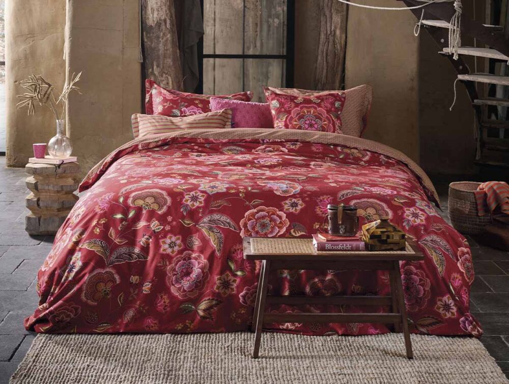 Ein gemütliches Schlafzimmer mit der Bettwäsche Pip Studio Matata mit rotem Blumenmuster, passender Bettdecke und Kissen, einer Bank mit Büchern und einer Tasse am Fußende des Bettes, einem gewebten Teppich, rustikalem Holzdekor und warmer, weicher Beleuchtung.