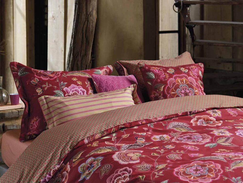 Das Bettwäsche Pip Studio Matata Set besteht aus einer roten Bettdecke mit Blumenmuster und passenden Kissen, die durch rosa, rote und grüne Streifen und geometrische Akzente in einem rustikalen Schlafzimmer hervorgehoben werden.