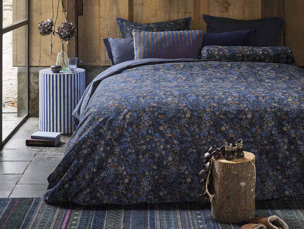 Ein gemütliches, rustikales Schlafzimmer mit einem Bett mit dunkelblauer, geblümter Bettwäsche von Pip Studio Tutti i Fiori, verschiedenen blauen Kissen, einem gestreiften Beistelltisch mit Büchern und Trockenblumen und einem Hocker mit Holzstumpf.