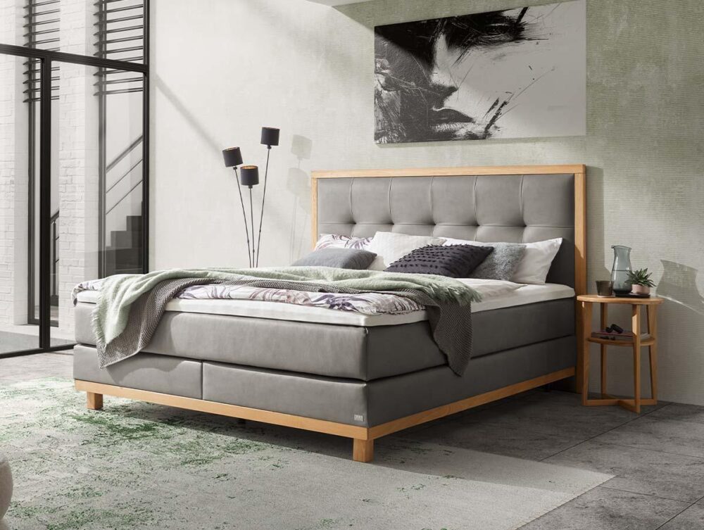 Modernes Schlafzimmer mit einem grau gepolsterten Bett, einem Holzrahmen und einem gepolsterten Kopfteil. Auf einem Beistelltisch stehen eine Vase und ein Glas. Abstrakte Kunst und eine hohe Lampe stehen im Hintergrund, während das Sonnenlicht durch große Fenster fällt.