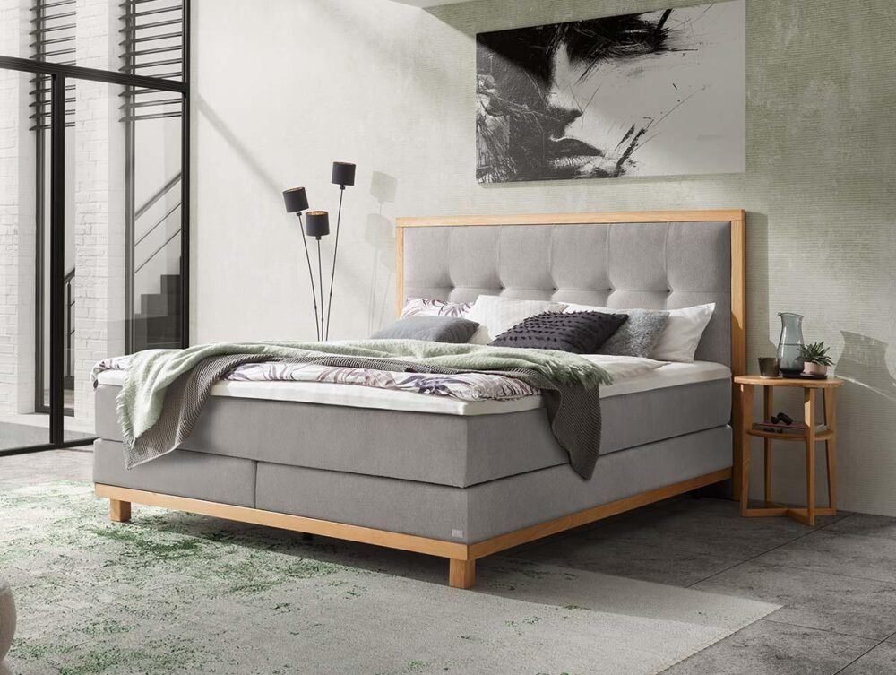 Ein modernes Schlafzimmer mit einem hellgrauen gepolsterten Bett, einer grünen Decke und einem kleinen hölzernen Nachttisch mit einer Glasvase. Über dem Bett hängt ein Schwarz-Weiß-Porträt, und die bodentiefen Fenster lassen natürliches Licht herein.