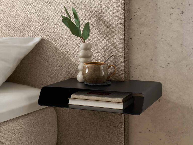Auf einem modernen, schwebenden schwarzen Nachttisch stehen eine gestapelte beigefarbene Vase mit grünen Blättern, eine braune Tasse auf einem Untersetzer und zwei Bücher neben einem beigen Bett und einer strukturierten Wand.