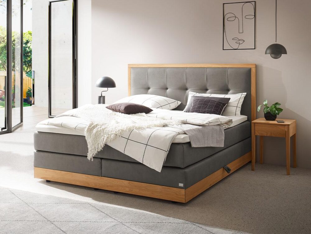 Ein modernes Schlafzimmer mit einem großen grauen Bett mit Holzrahmen, einem grauen getufteten Kopfteil, weißem und grauem Bettzeug, einem hölzernen Nachttisch mit einer Pflanze, einer Hängelampe und minimalistischer Linienkunst an der Wand. Sonnenlicht füllt den Raum.