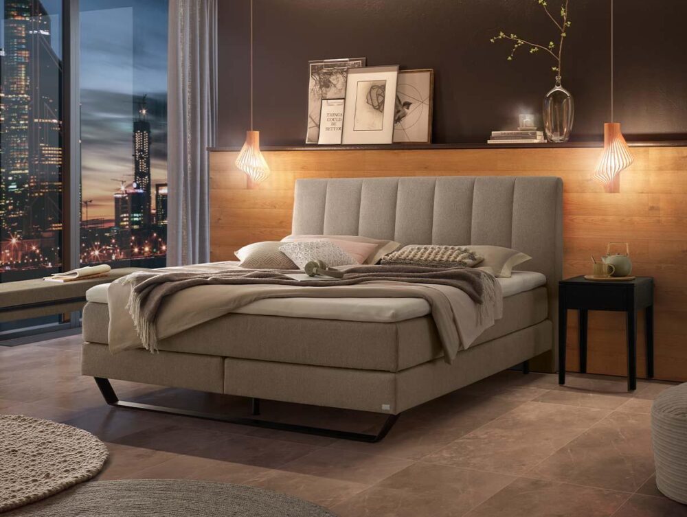 Ein modernes Schlafzimmer verfügt über ein beigefarbenes Polsterbett mit kuscheliger Bettwäsche, Pendelleuchten, einen schwarzen Nachttisch und dekorative Kunst an einer Akzentwand aus Holz. Vom Boden bis zur Decke reichende Fenster zeigen die Skyline der Stadt in der Abenddämmerung.