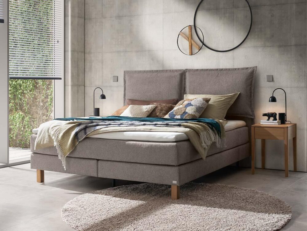 Ein modernes Schlafzimmer mit einem beigen Polsterbett, verschiedenen Kissen und einer blauen Überwurfdecke. Es gibt zwei hölzerne Nachttische mit schwarzen Lampen, einen runden Spiegel an der Wand und einen runden beigen Teppich auf einem hellen Fliesenboden.