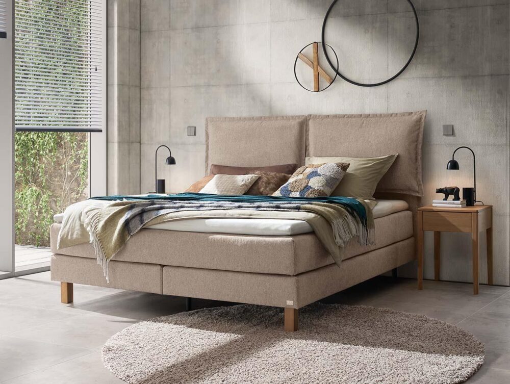 Ein modernes Schlafzimmer mit einem beigen Polsterbett, verschiedenen Kissen und einer Decke, einem hölzernen Nachttisch mit einer Lampe und einer minimalistischen Einrichtung. Das Sonnenlicht fällt durch große Fenster mit Jalousien. Ein runder Teppich liegt auf dem Boden.