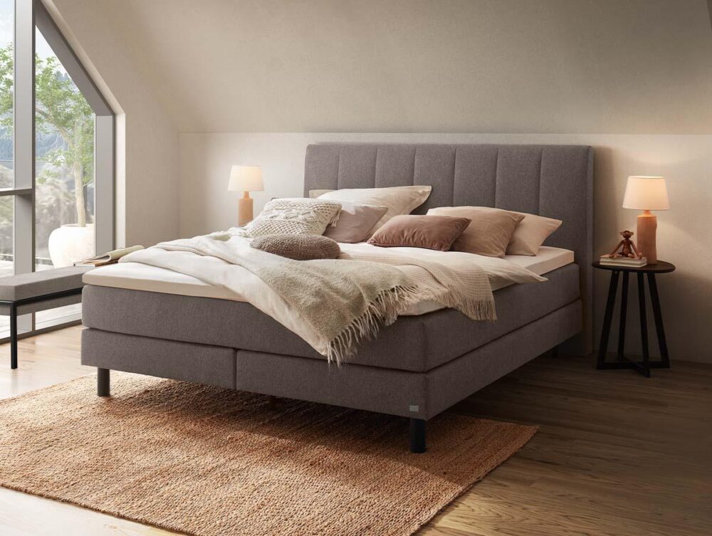 Ein gemütliches Schlafzimmer mit einem großen, gepolsterten Bett, das mit Kissen und Decken in neutralen Farbtönen bezogen ist, einem gewebten Teppich, zwei Nachttischen mit Lampen und einem Fenster, das natürliches Licht hereinlässt.