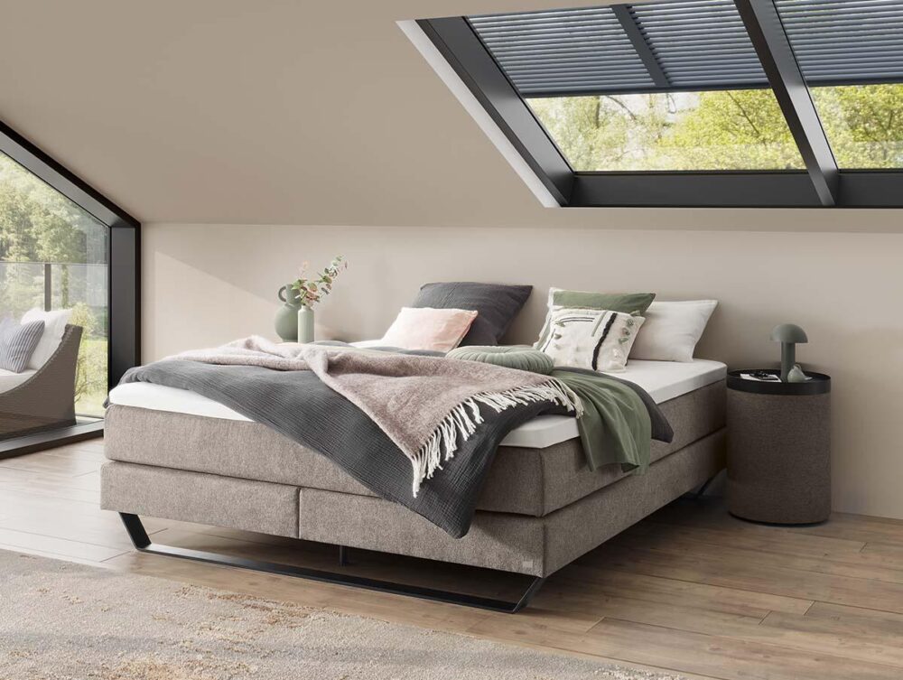 Ein modernes Schlafzimmer mit einem beigen Polsterbett, das mit Kissen und einer Decke ausgelegt ist, befindet sich unter einem großen Dachfenster. Natürliches Licht durchflutet den Raum und unterstreicht die Holzböden und die minimalistische Einrichtung.