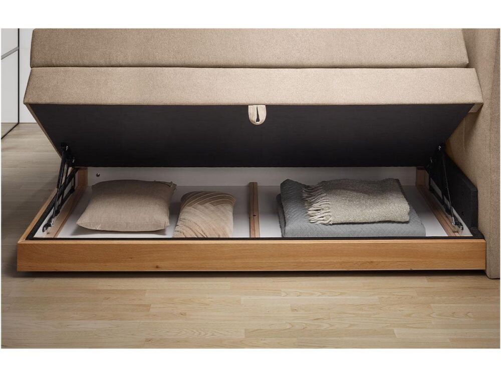 Ein beigefarbenes Sofa, dessen Sitz hochgeklappt ist, so dass ein Staufach darunter zum Vorschein kommt, in dem sich ein Kissen, ein Polster und eine gefaltete Decke befinden. Das Ablagefach hat einen Holzrahmen und steht auf einem hellen Holzboden.