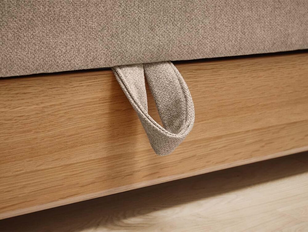 Nahaufnahme eines Schlaufengriffs aus Stoff, der an der Seite eines beigefarbenen gepolsterten Bettrahmens über einem hölzernen Bettgestell befestigt ist. Das Bett steht auf einem hellen Boden.