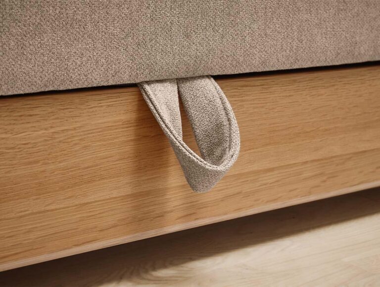 Nahaufnahme eines Schlaufengriffs aus Stoff, der an der Seite eines beigefarbenen gepolsterten Bettrahmens über einem hölzernen Bettgestell befestigt ist. Das Bett steht auf einem hellen Boden.