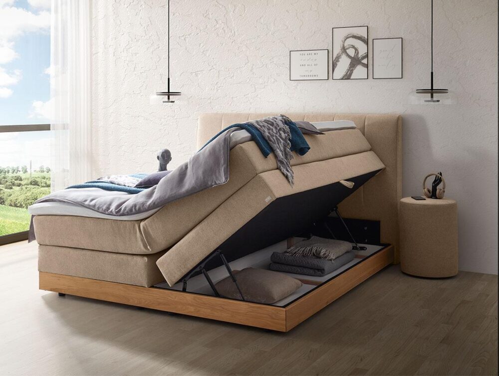 Ein beiges Stauraumbett mit hochgeklappter Matratze, unter der sich ein Stauraum mit Bettwäsche befindet. Das Zimmer ist modern eingerichtet, hat ein großes Fenster mit Blick auf die Landschaft und minimalistische Kunstwerke an der Wand.