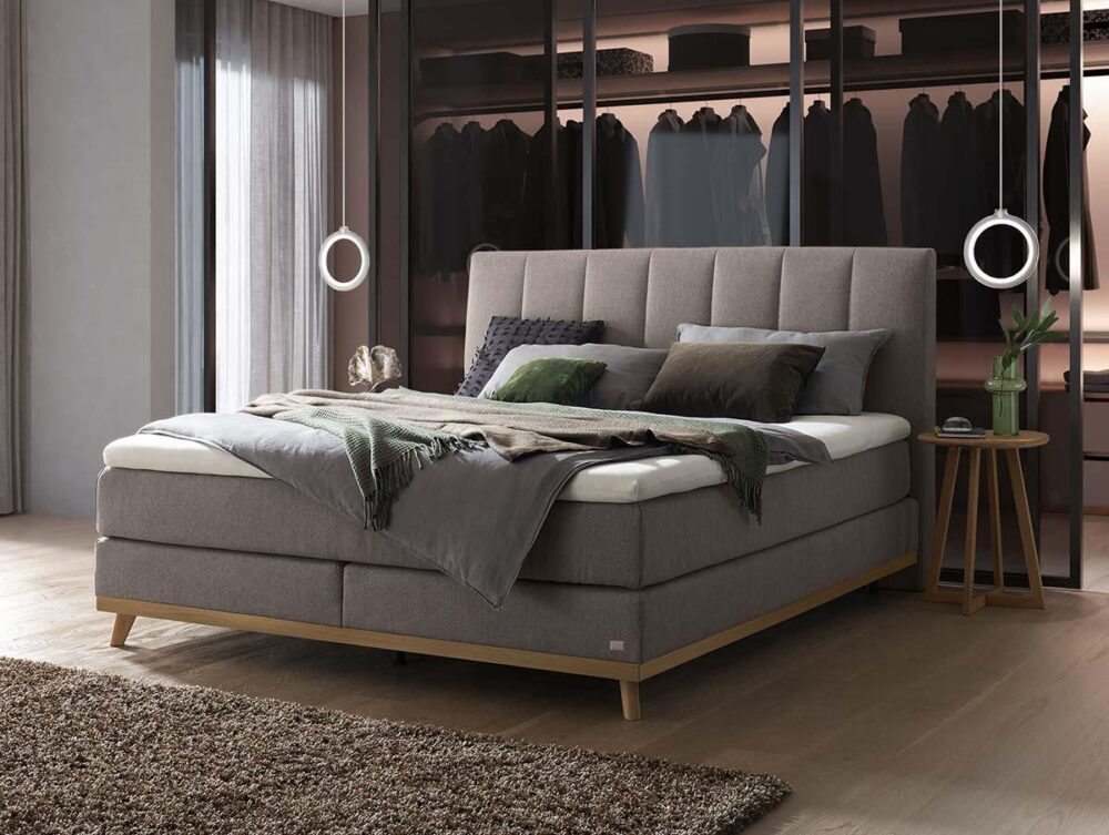 Ein modernes Schlafzimmer mit einem großen, gepolsterten Bett in Grautönen, das mit Kissen und Decken ausgelegt ist. Hinter dem Bett befindet sich ein offener Schrank mit hängenden Kleidungsstücken, Regalen und sanfter Beleuchtung. Zwei Pendelleuchten hängen an beiden Seiten des Bettes.